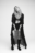 AI-generated fashion portrait of a woman in a long sheer coat, layered necklaces, and boots, striking a confident pose against a blank background
