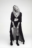 AI-generated fashion portrait of a woman in a long sheer coat, layered necklaces, and boots, striking a confident pose against a blank background