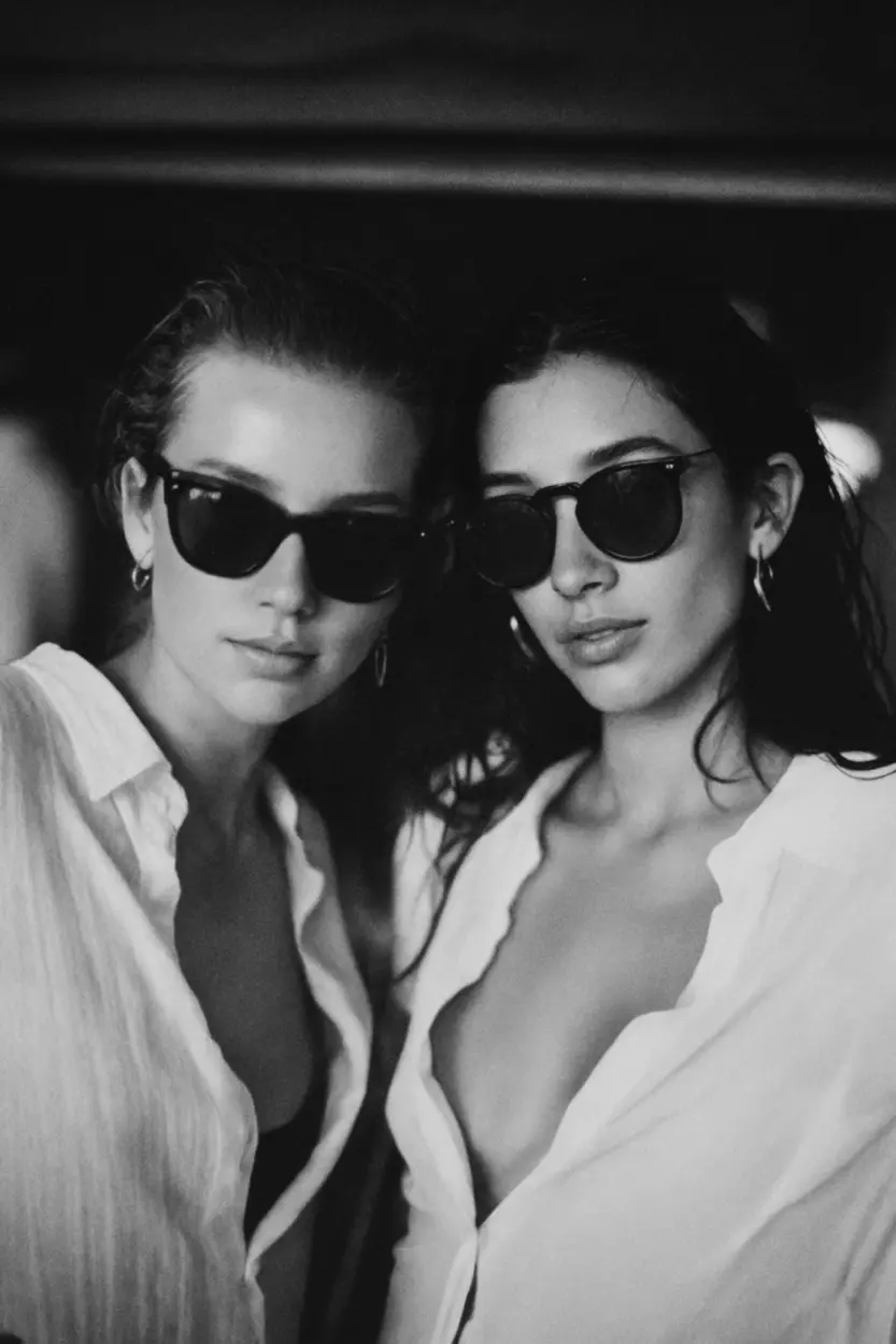 Black and white photo of two women wearing Ray Ban sunglasses and open white shirts, styled like a film still from a fashion-forward AI shoot